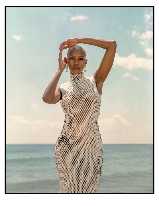 A woman with short blonde hair poses in a mesh dress with her arms raised above her head, standing in front of the ocean under a cloudy sky.