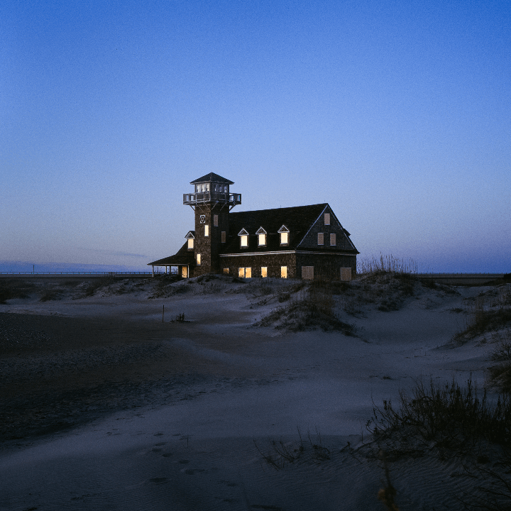 A building with a tower and illuminated windows stands on a sandy dune under a clear evening sky.