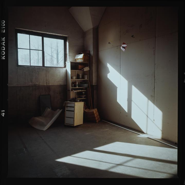 Sunlight streams through a window into a sparsely furnished room with concrete floors and a wooden shelf.