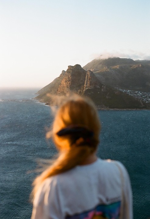 A woman with a ponytail looks out at a mountainous coastline with a town nestled at its base, under a hazy sky.