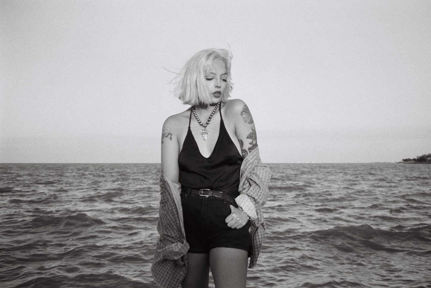 A woman with short blonde hair and tattoos stands in front of a body of water.