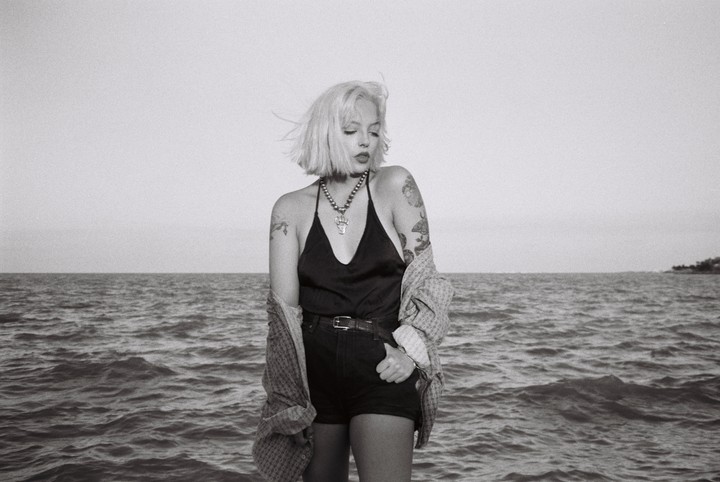 A woman with short blonde hair and tattoos stands in front of a body of water.