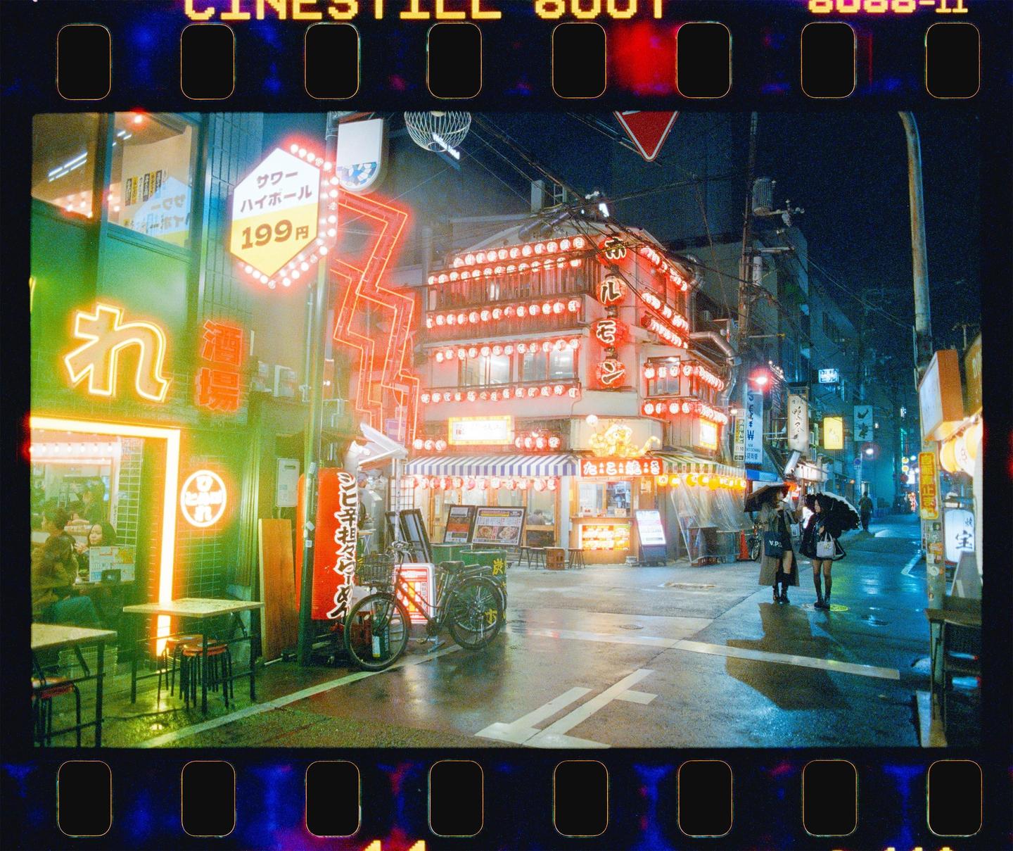 Two women with umbrellas walk down a neon-lit street at night.