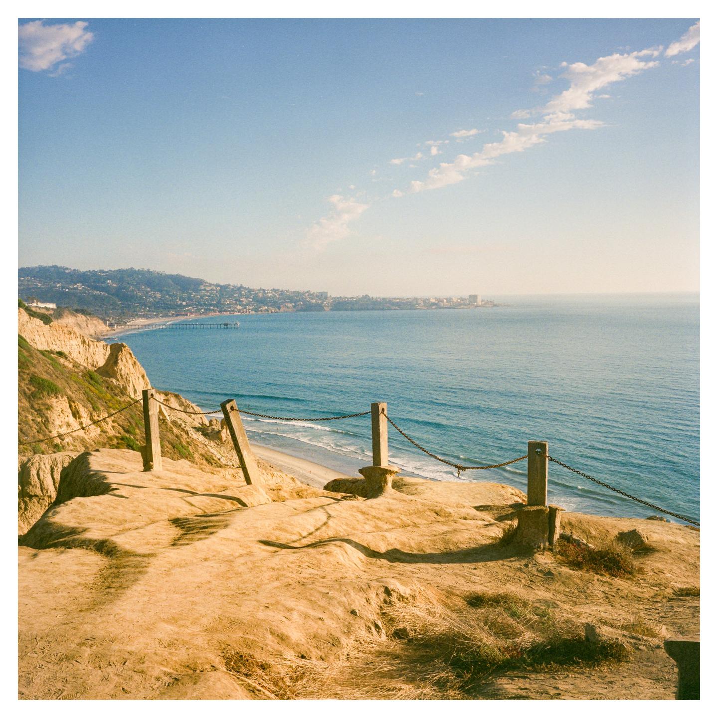A cliff overlooks a beach and ocean with a distant city under a blue sky.