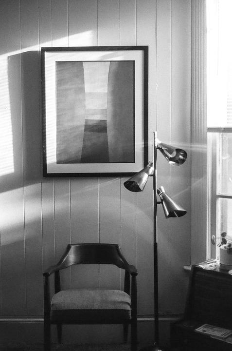 Sunlight streams through a window, illuminating a chair, a floor lamp, and a framed picture on a wall.