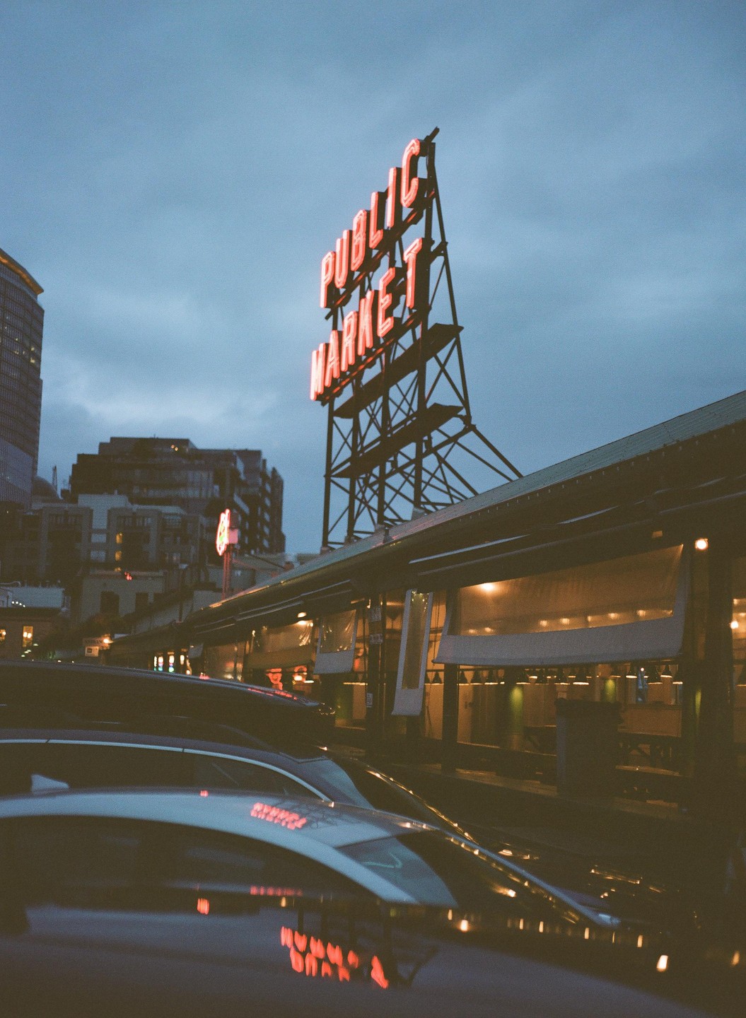 A neon sign for a public market stands tall against a cloudy evening sky, with cars and buildings visible below.