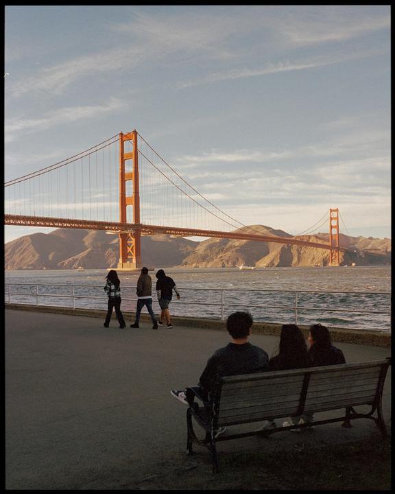 Three people sit on a bench looking at the Golden Gate Bridge while three other people walk along the water.