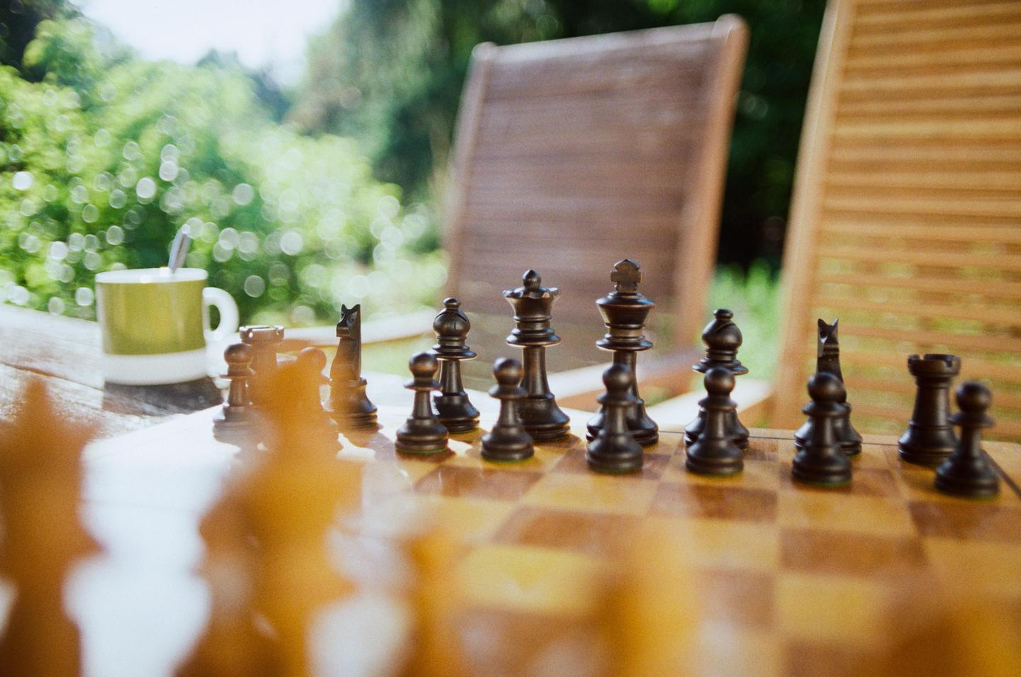A chessboard with black pieces is set up on a wooden table next to a green mug, with green foliage and wooden chairs in the background.