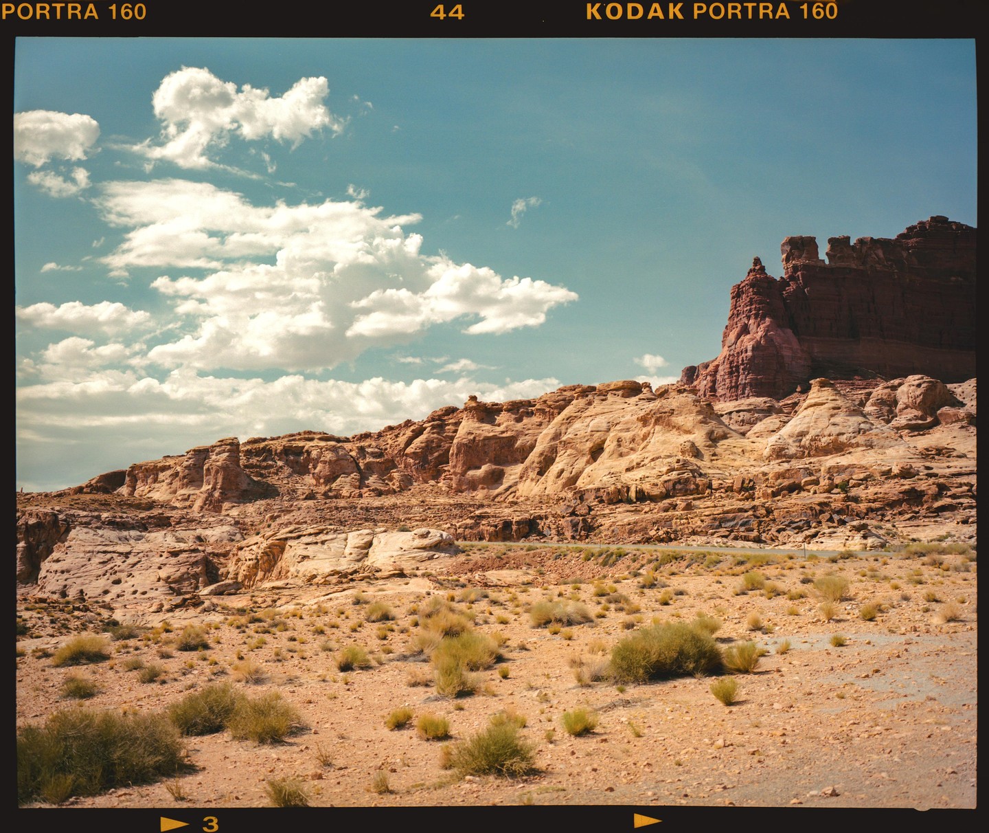 A desert landscape features a road winding through rocky terrain with sparse vegetation under a blue sky with clouds.