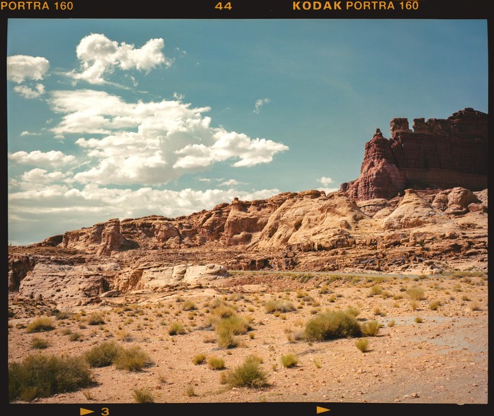 A desert landscape features a road winding through rocky terrain with sparse vegetation under a blue sky with clouds.