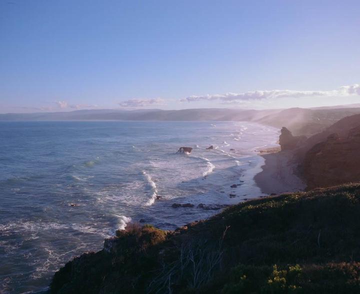 A wide view of the Australian coastline shows waves crashing on a beach next to cliffs and rock formations.