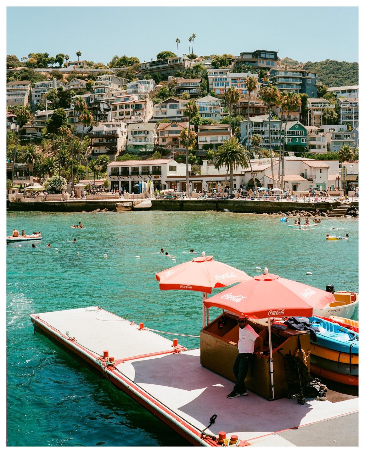 A man stands under two red umbrellas on a dock with a hillside town and ocean in the background.