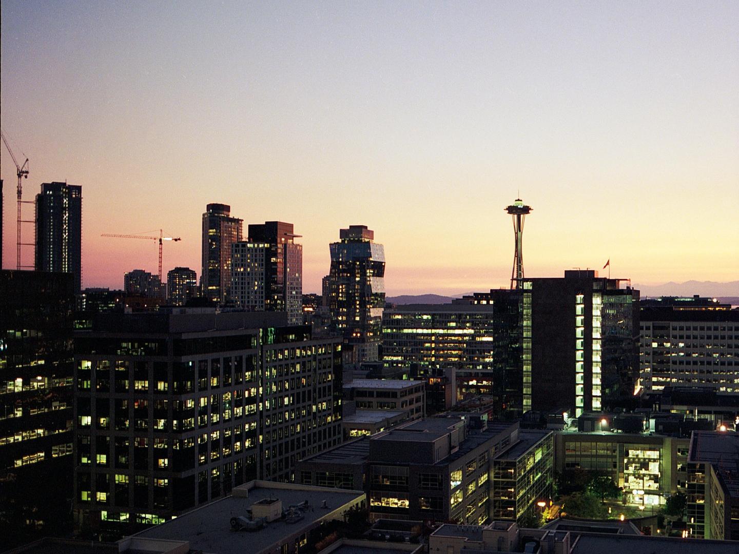 A cityscape of Seattle at dusk shows the Space Needle and other buildings with illuminated windows against a colorful sky.