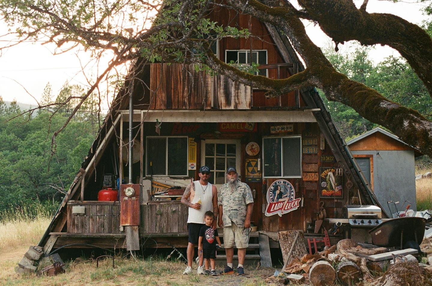 A man, a boy, and another man stand in front of a rustic wooden cabin with a large tree branch overhead.