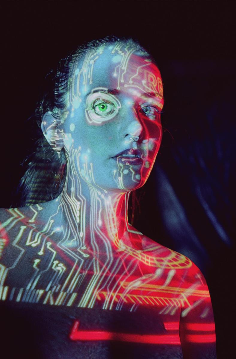 A woman's face and upper body are illuminated by projected circuit board patterns in red and blue light.
