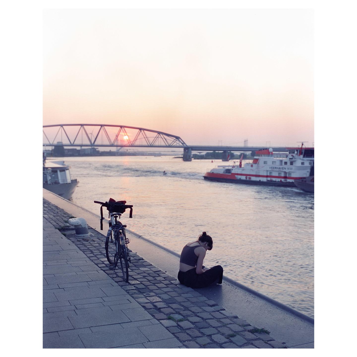 A woman sits on a cobblestone riverbank next to a bicycle, with a bridge and boats in the background at sunset.