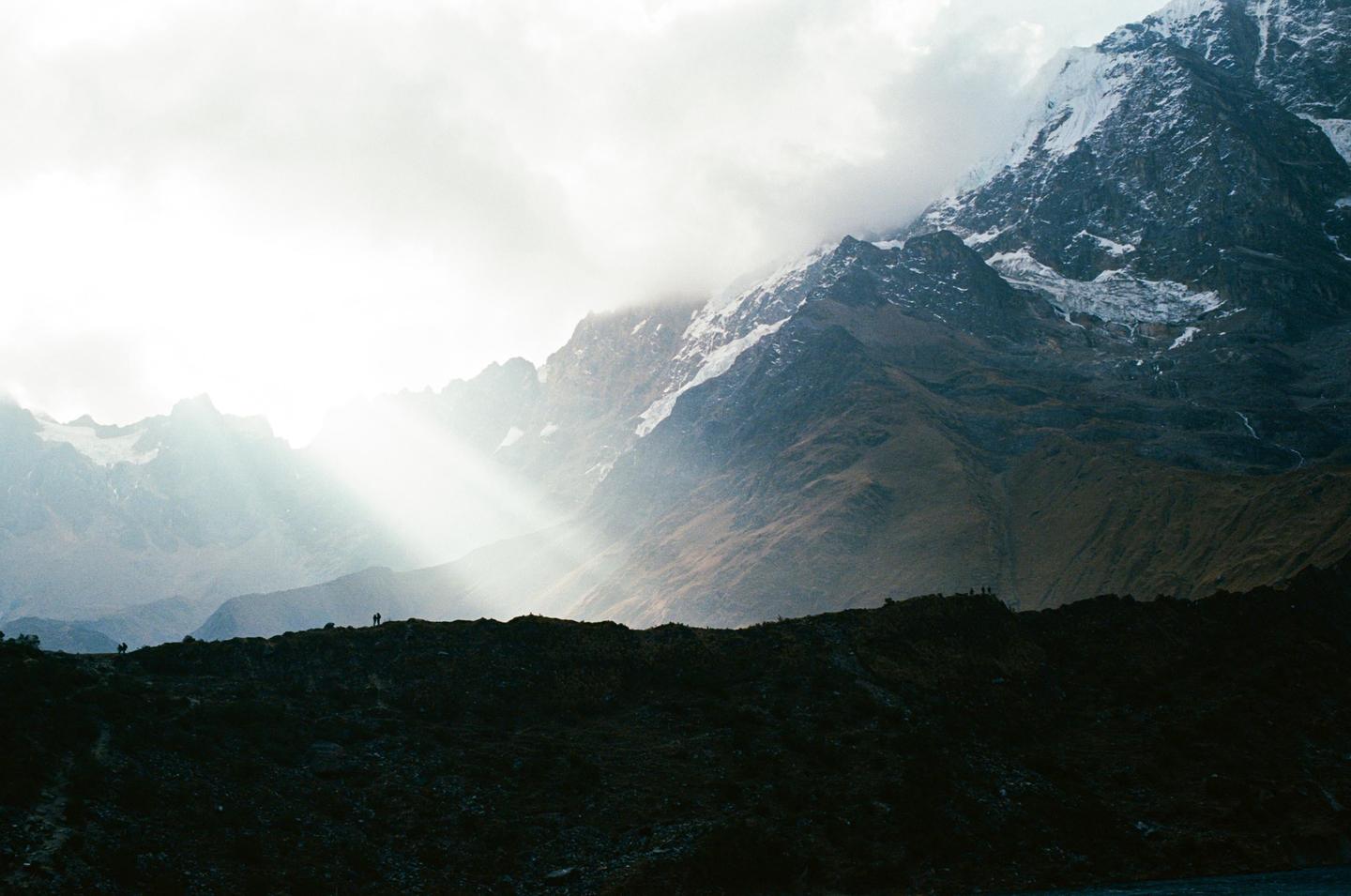 Sunlight breaks through clouds over a mountain range with snow-capped peaks and a dark ridge in the foreground.
