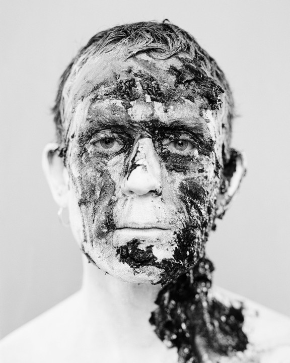 A man with dark and light paint covering his face and neck looks directly forward.