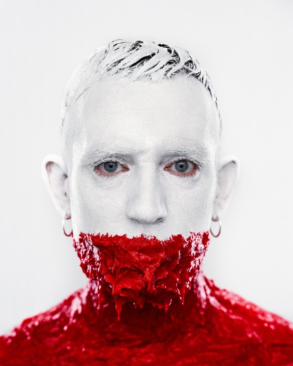 A man with white paint on his face and red paint dripping from his mouth and neck stares forward.