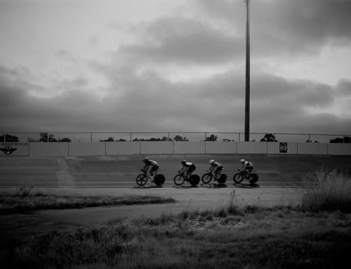 Four cyclists race on a track under a cloudy sky.