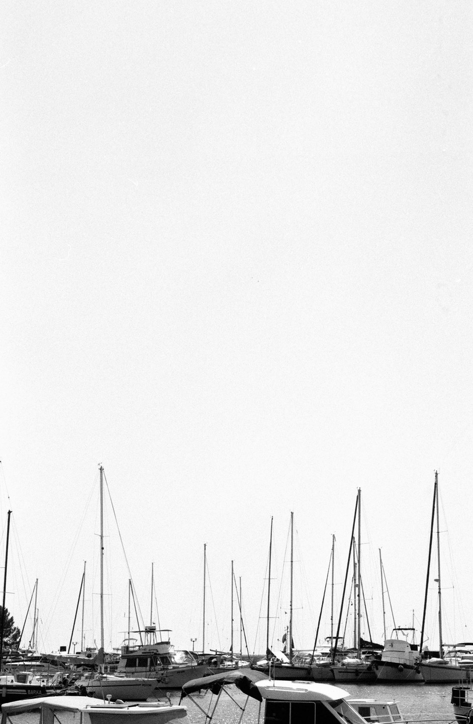 A collection of boats with tall masts are docked in a harbor under a bright sky.