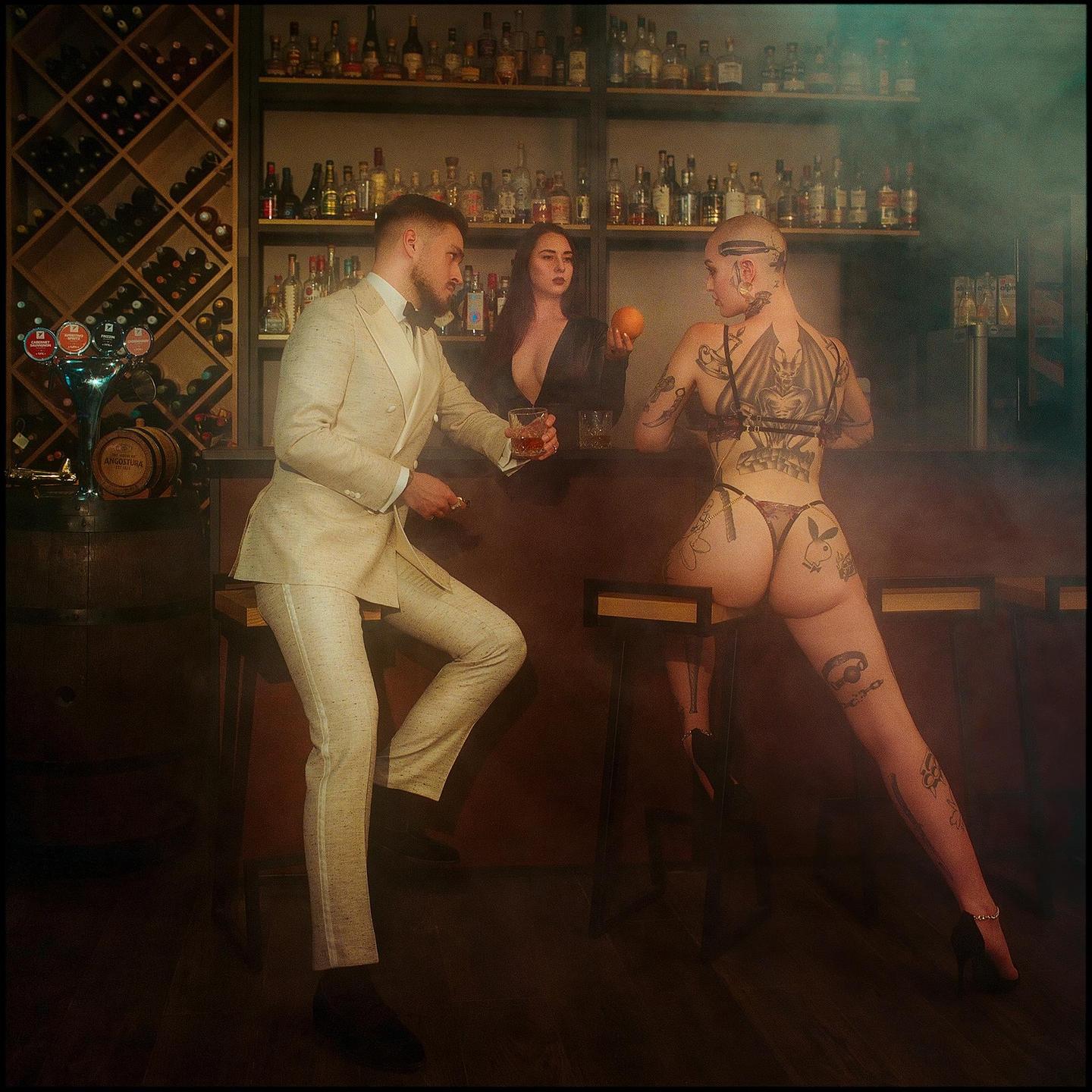 A man in a suit sits at a bar with two women, one of whom is holding an orange.