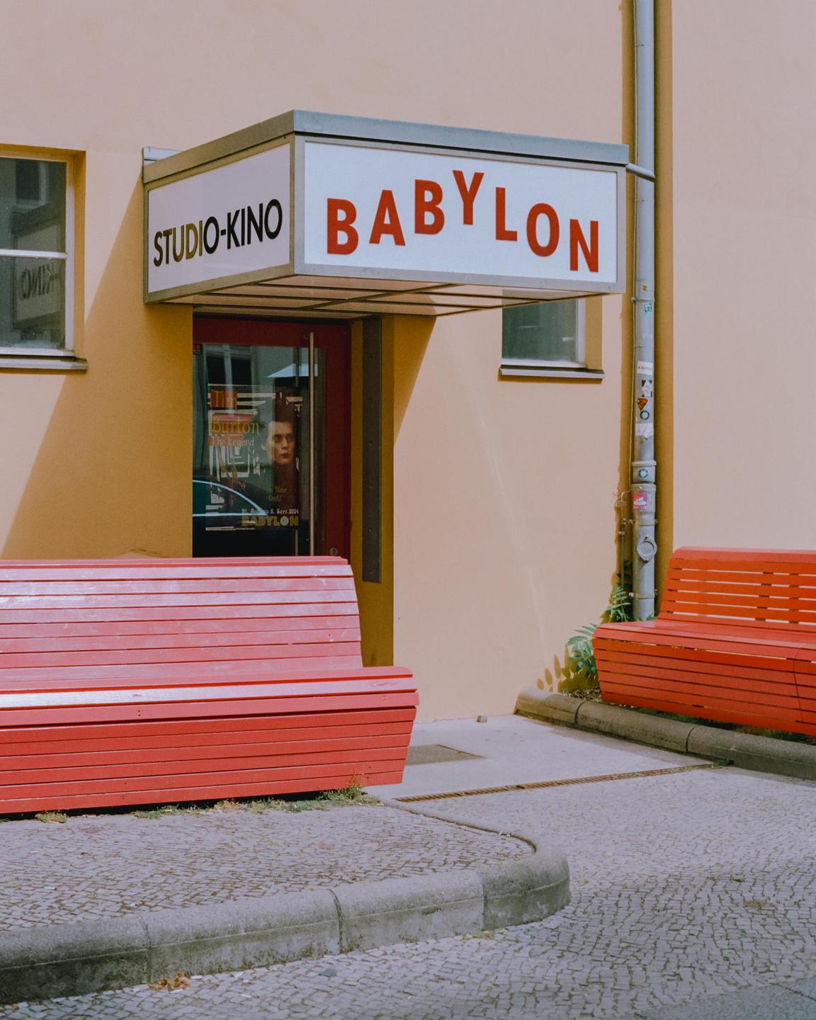 Two red benches sit on a cobblestone sidewalk in front of a cinema entrance with a "Babylon" sign.