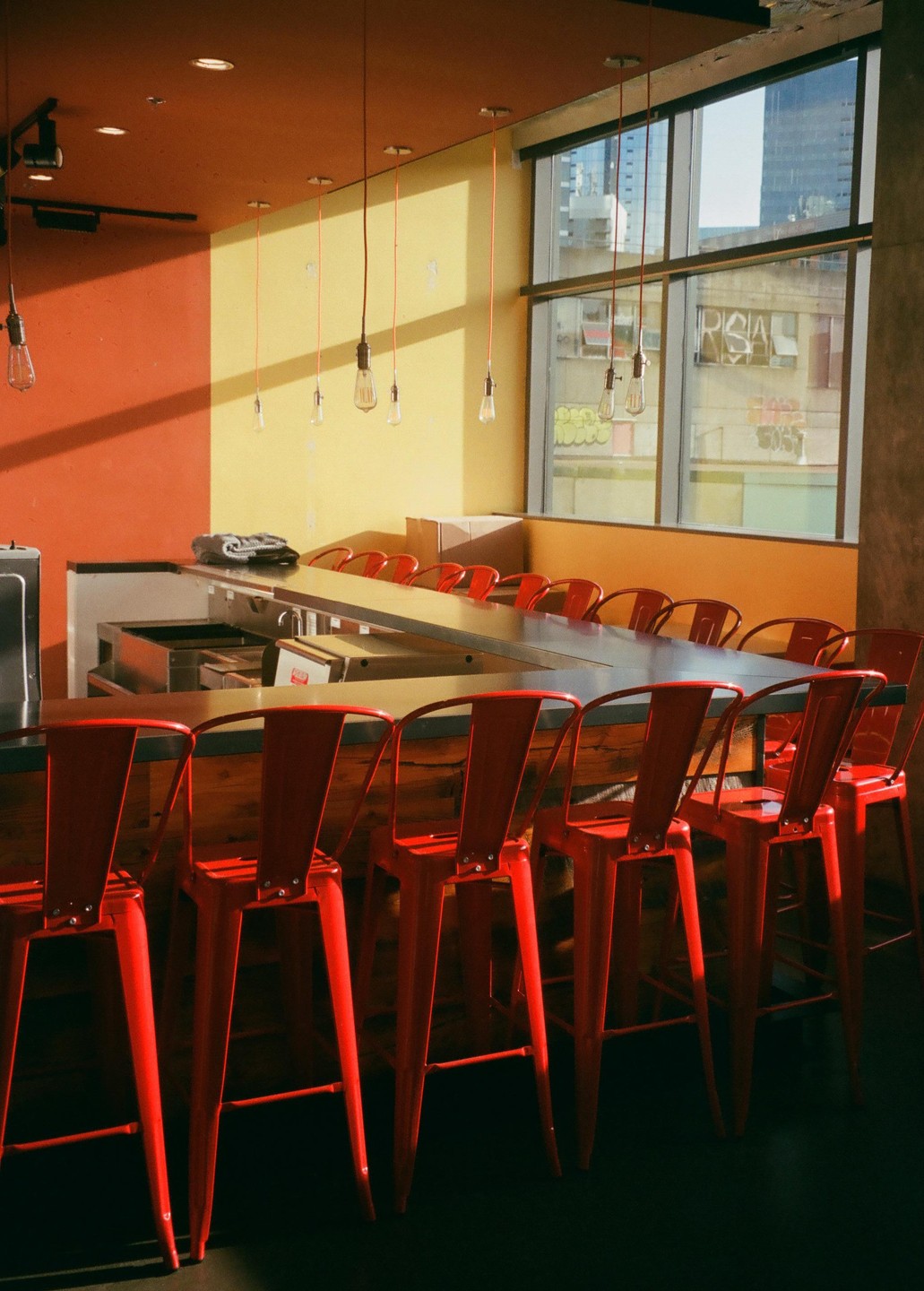 A row of red bar stools sits in front of a counter with a large window looking out onto a city.