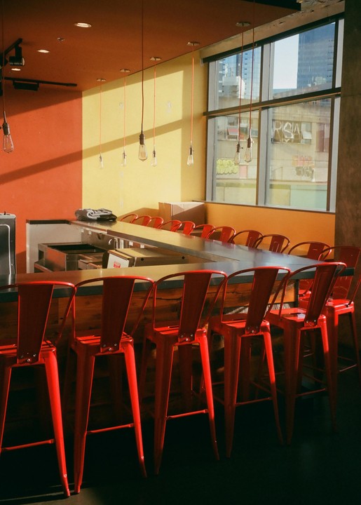 A row of red bar stools sits in front of a counter with a large window looking out onto a city.