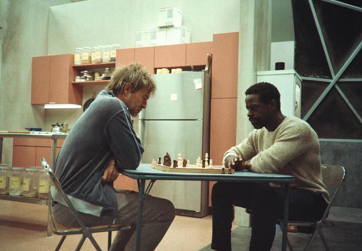 Two men sit across from each other at a table with a chessboard between them in a kitchen.