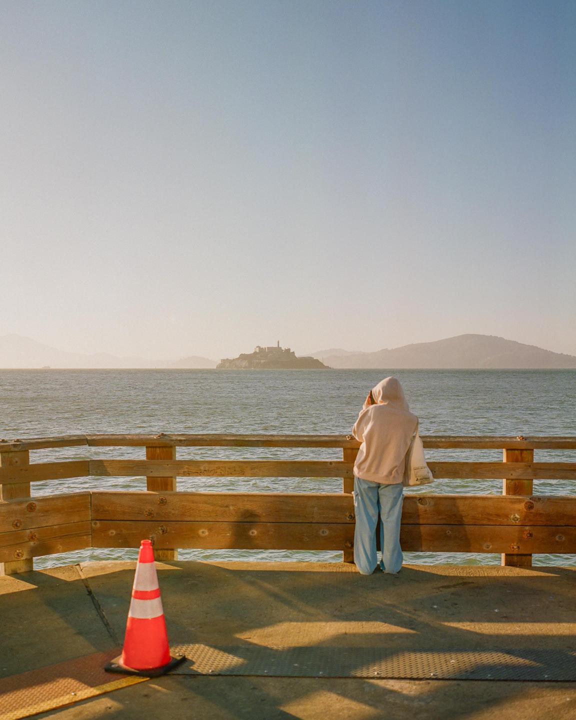 A person in a hoodie stands on a pier looking out at Alcatraz Island in the distance during golden hour.
