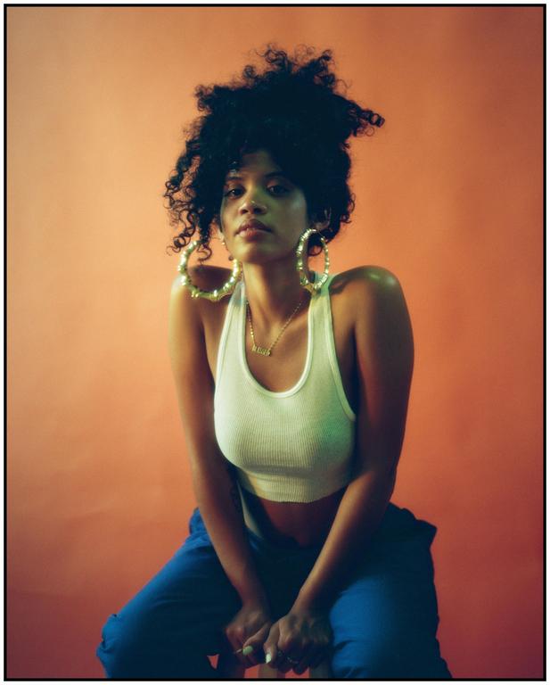 A woman with curly hair and large hoop earrings sits against an orange background.