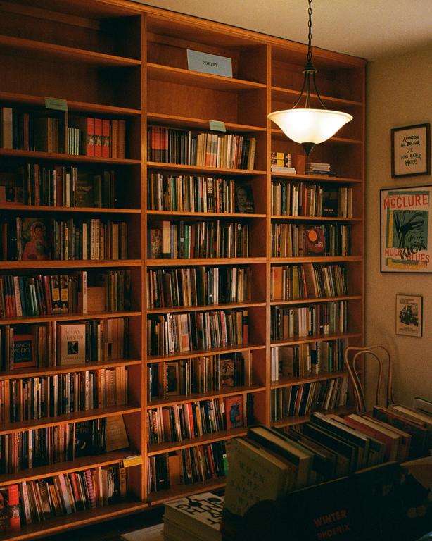 A large wooden bookshelf filled with books stands in a room with a hanging lamp and framed pictures on the wall.