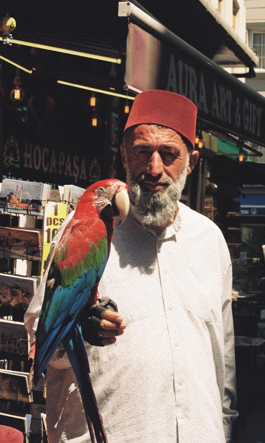 A man wearing a red fez and a white shirt holds a colorful parrot on his gloved hand.