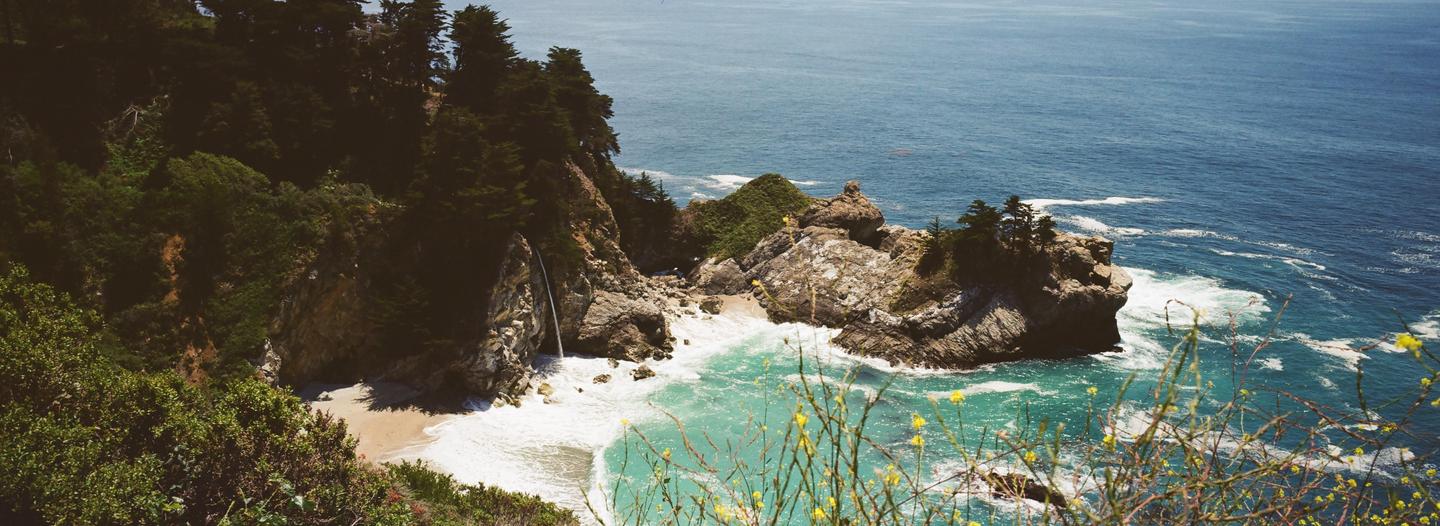 A waterfall cascades onto a sandy beach next to the ocean, with green foliage and rocks surrounding the scene.