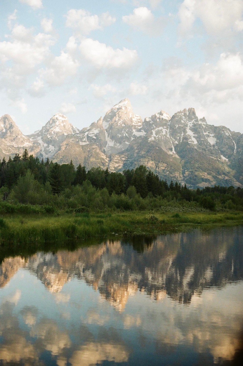Snow-capped mountains rise above a forest and are reflected in a calm body of water under a cloudy sky.