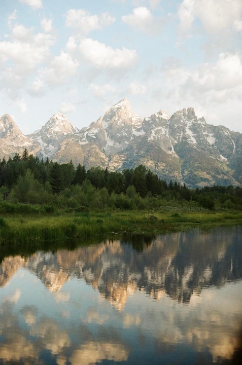 Snow-capped mountains rise above a forest and are reflected in a calm body of water under a cloudy sky.