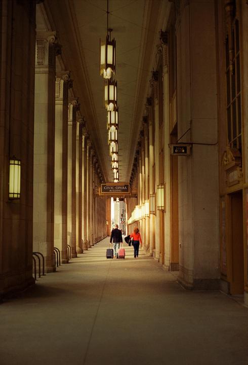 A man and a woman walk with luggage down a long, columned hallway with hanging lights.