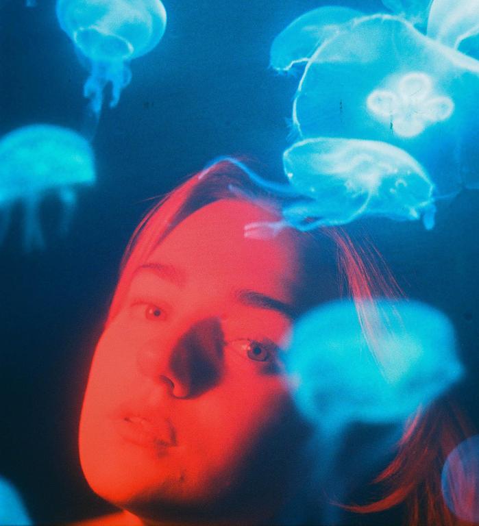 A woman's face is illuminated by red light while blue jellyfish float around her.