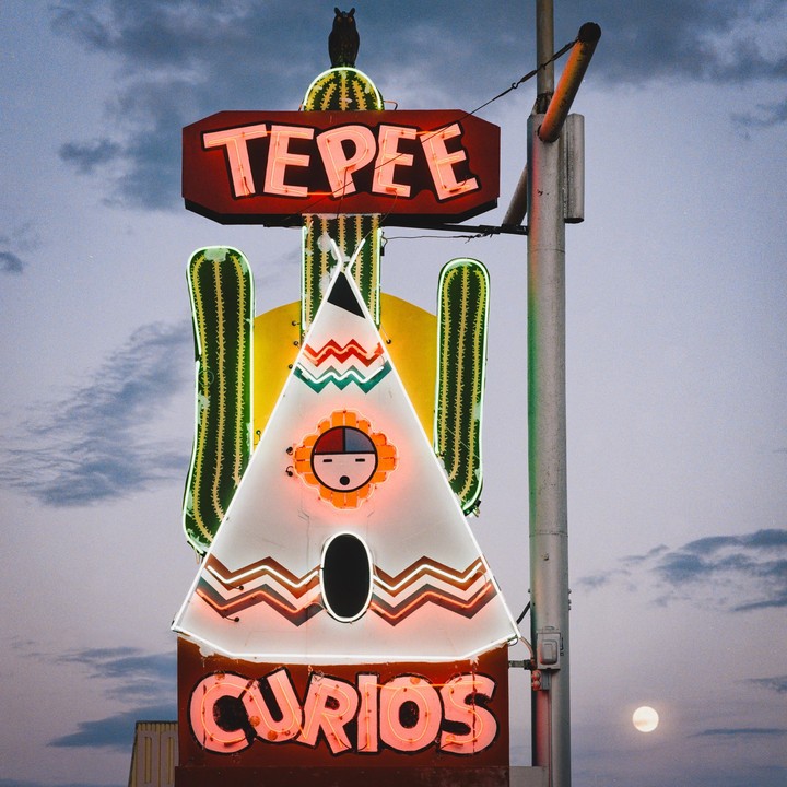 A neon sign shaped like a teepee and saguaro cactus stands against a cloudy sky with a full moon visible.