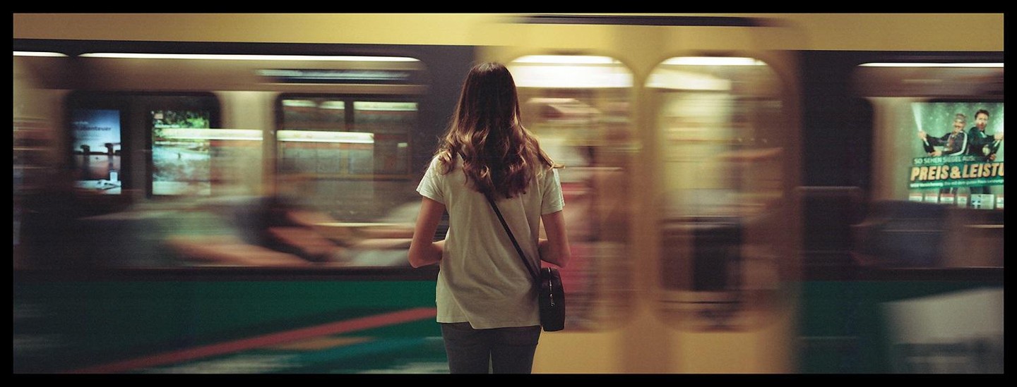 A woman stands on a subway platform as a train passes by, creating motion blur.