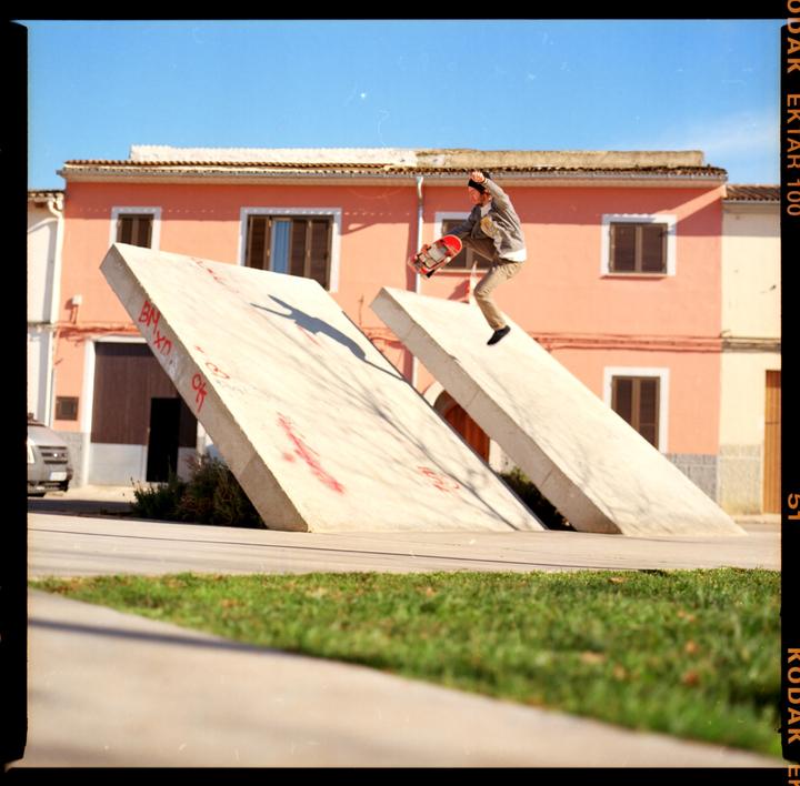 A skateboarder jumps in the air over a concrete ramp in front of a pink building.