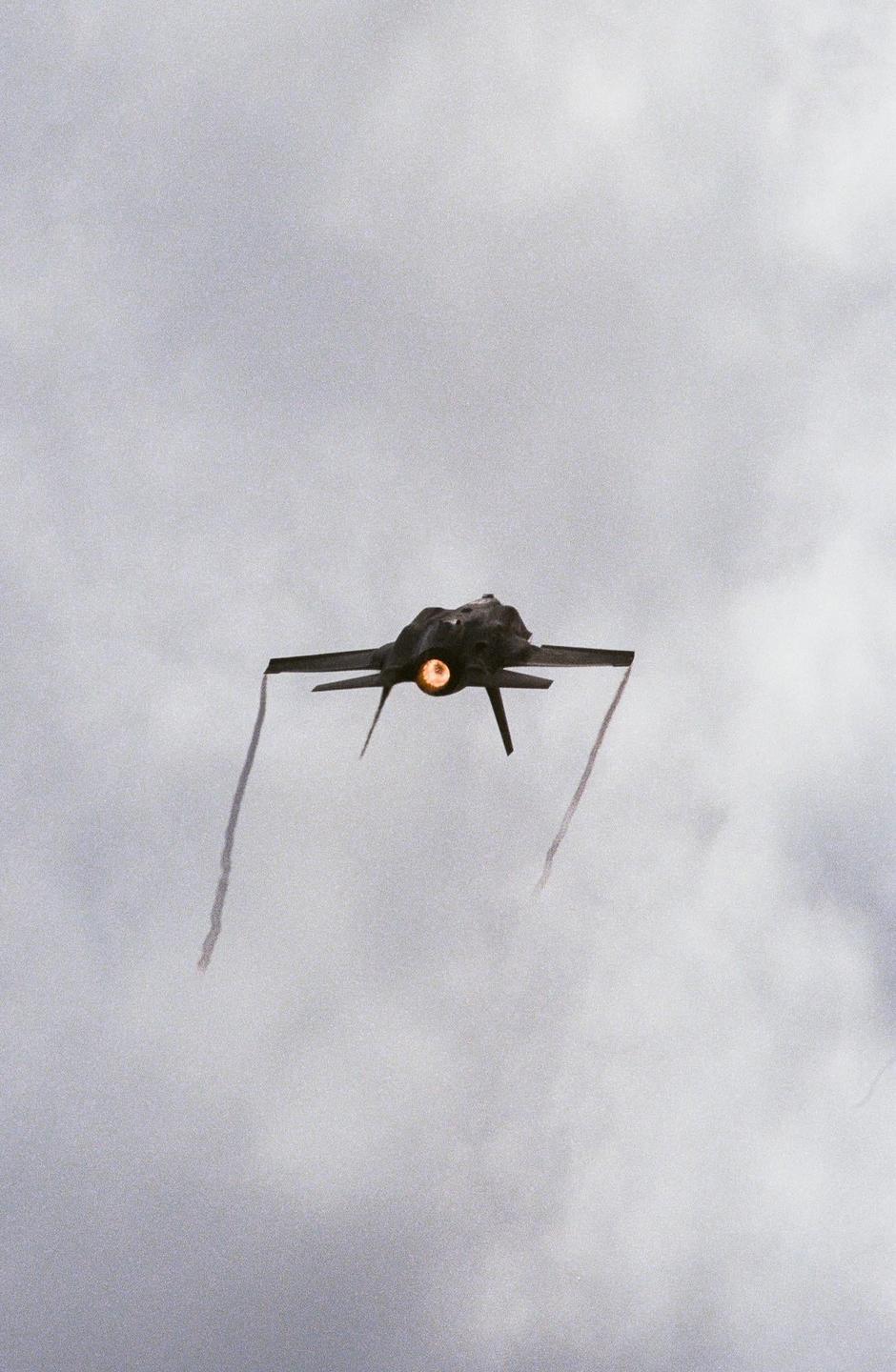 A jet aircraft with an illuminated engine exhaust flies through a cloudy sky, leaving two trails of smoke.
