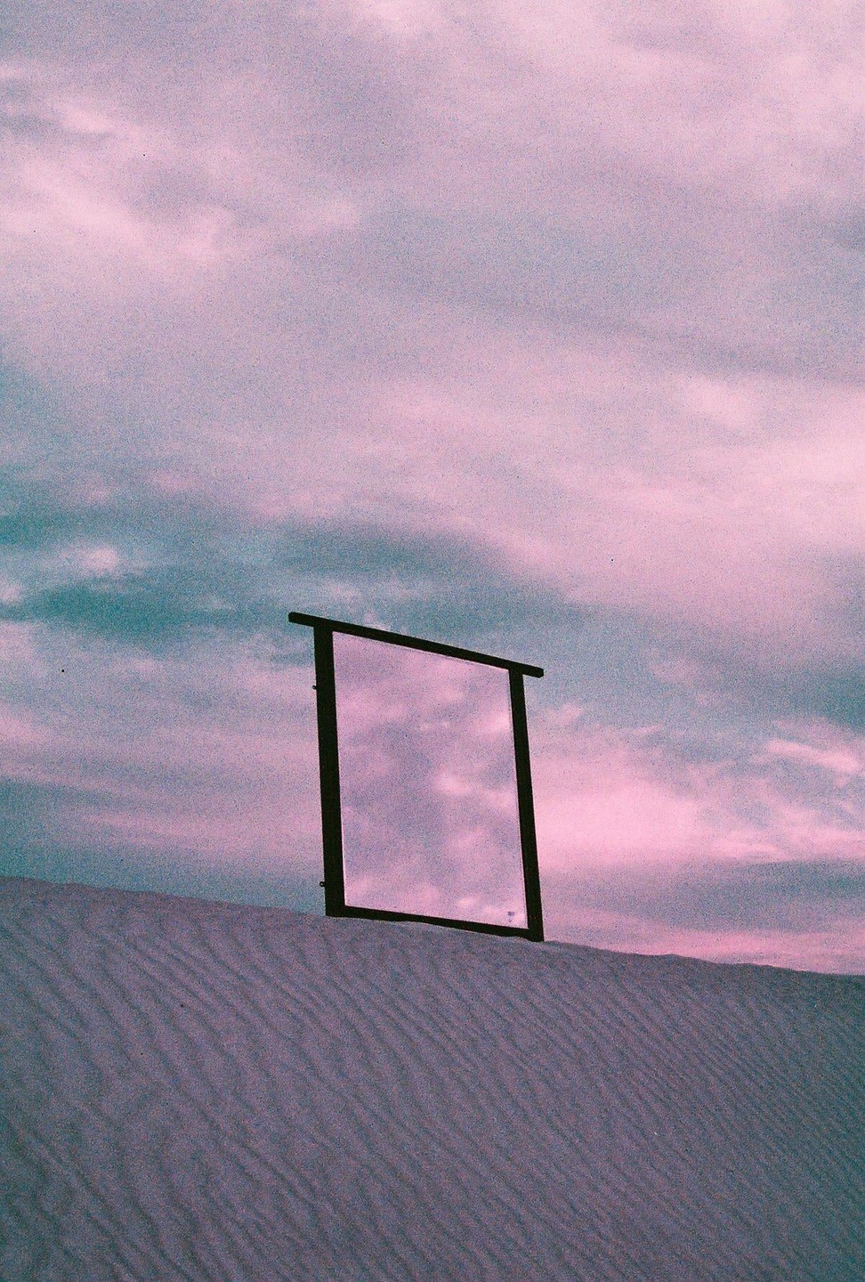 A mirror stands in sand dunes reflecting a purple and blue sky.