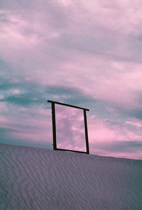 A mirror stands in sand dunes reflecting a purple and blue sky.