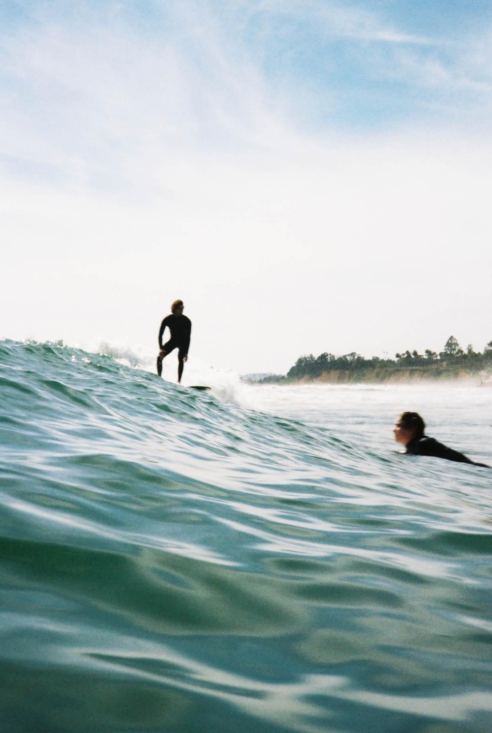 A surfer rides a wave while another surfer paddles in the water nearby.