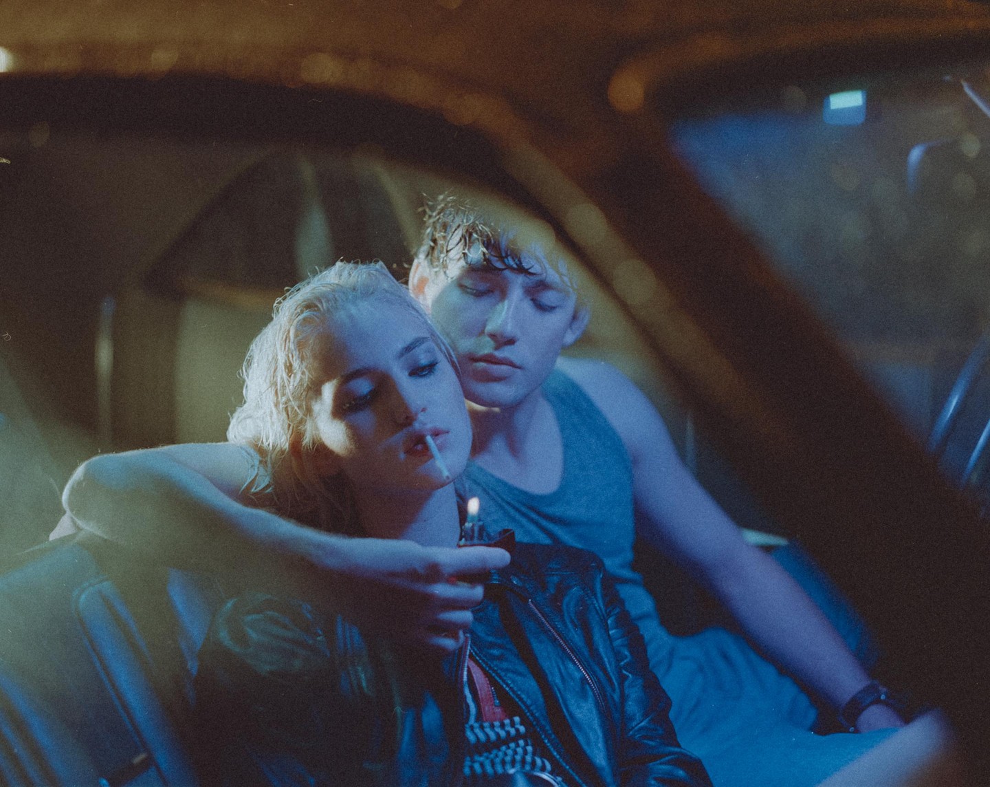 A couple sits in a car at night, and the woman lights a cigarette.
