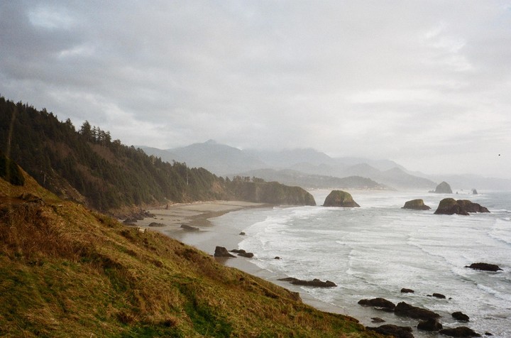 A wide view of the Oregon coast shows a forested hillside overlooking a beach with sea stacks and mountains in the distance under a cloudy sky.
