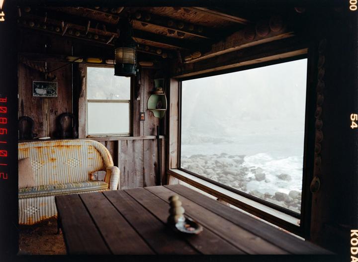 A rustic wooden room with a large window overlooks a rocky, misty coastline.