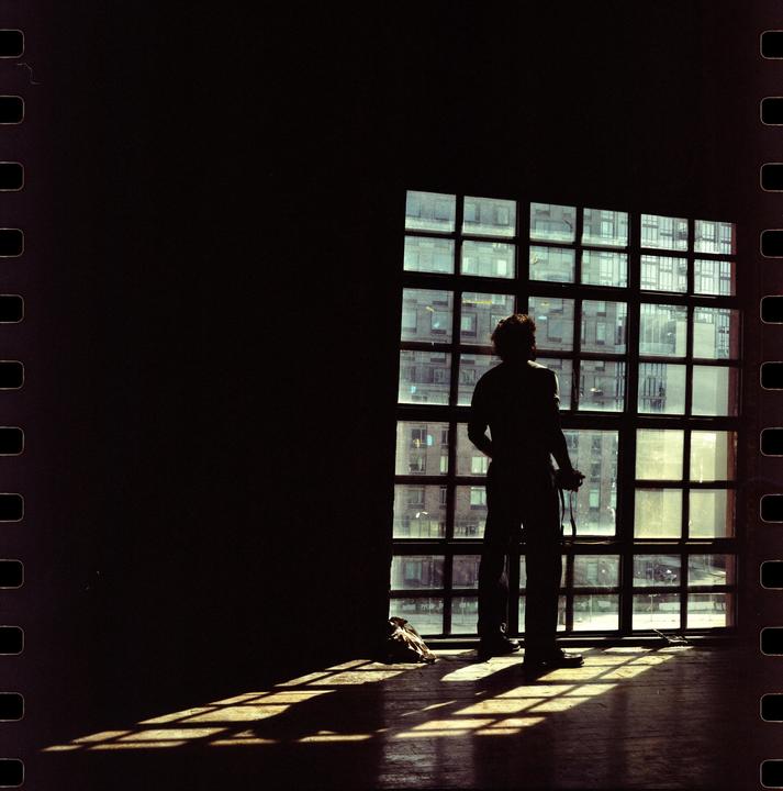 A man stands silhouetted against a large grid window, looking out at city buildings.
